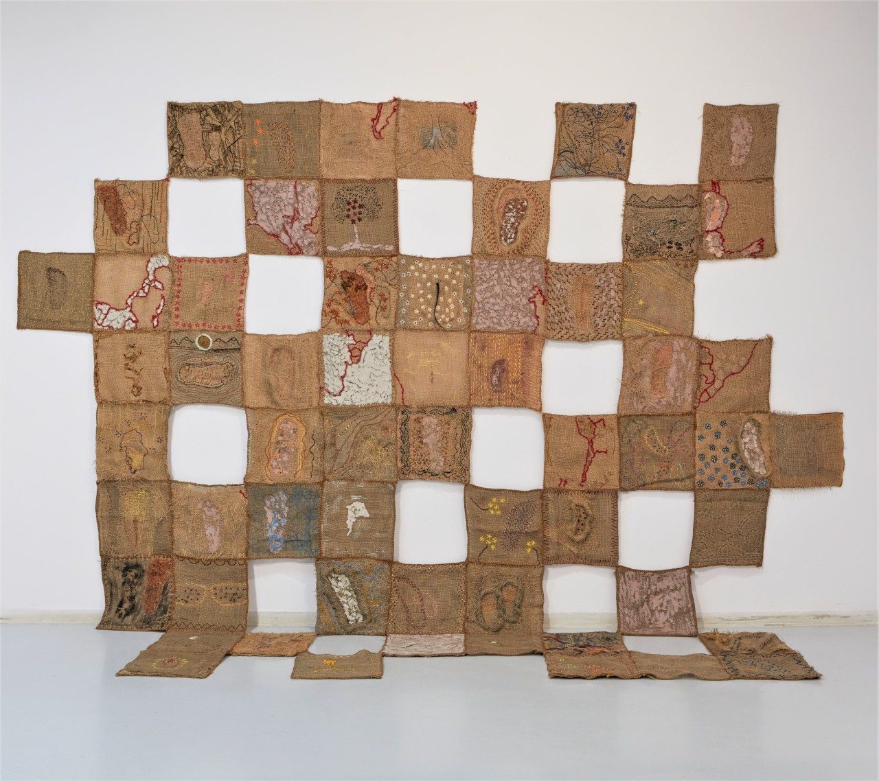 Hanging embroidered patchwork piece made out of jute, assembled in a checker pattern with empty spaces.