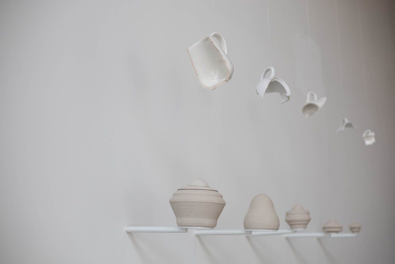 suspended fragments of ceramic cups and containers, under the suspended ceramics 3D modeled copy of the object in movement sit on a minimalist on a shelf fixed on a grey wall