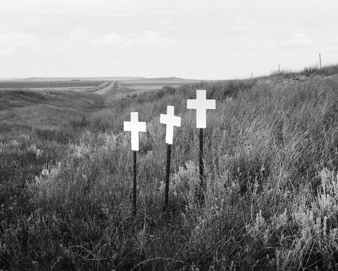 Black and white photograph depicting a rural road and three white crosses planted in the grass as markers of deadly road accidents.