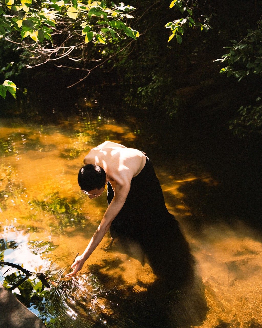 photographic portrait in colour of young person of vietnamese descent standing in a river and gently touching the water on a warm summer day