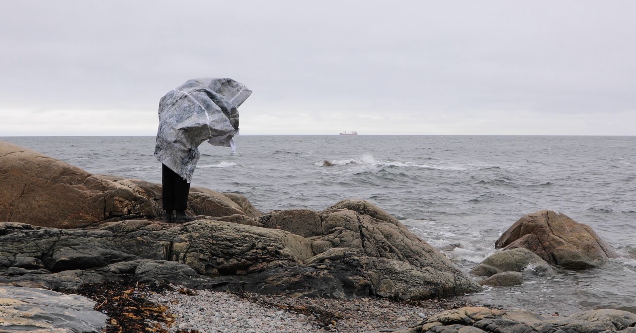 female artist standing on rocks facing the ocean, drapped in a large piece of paper with a rock motif 