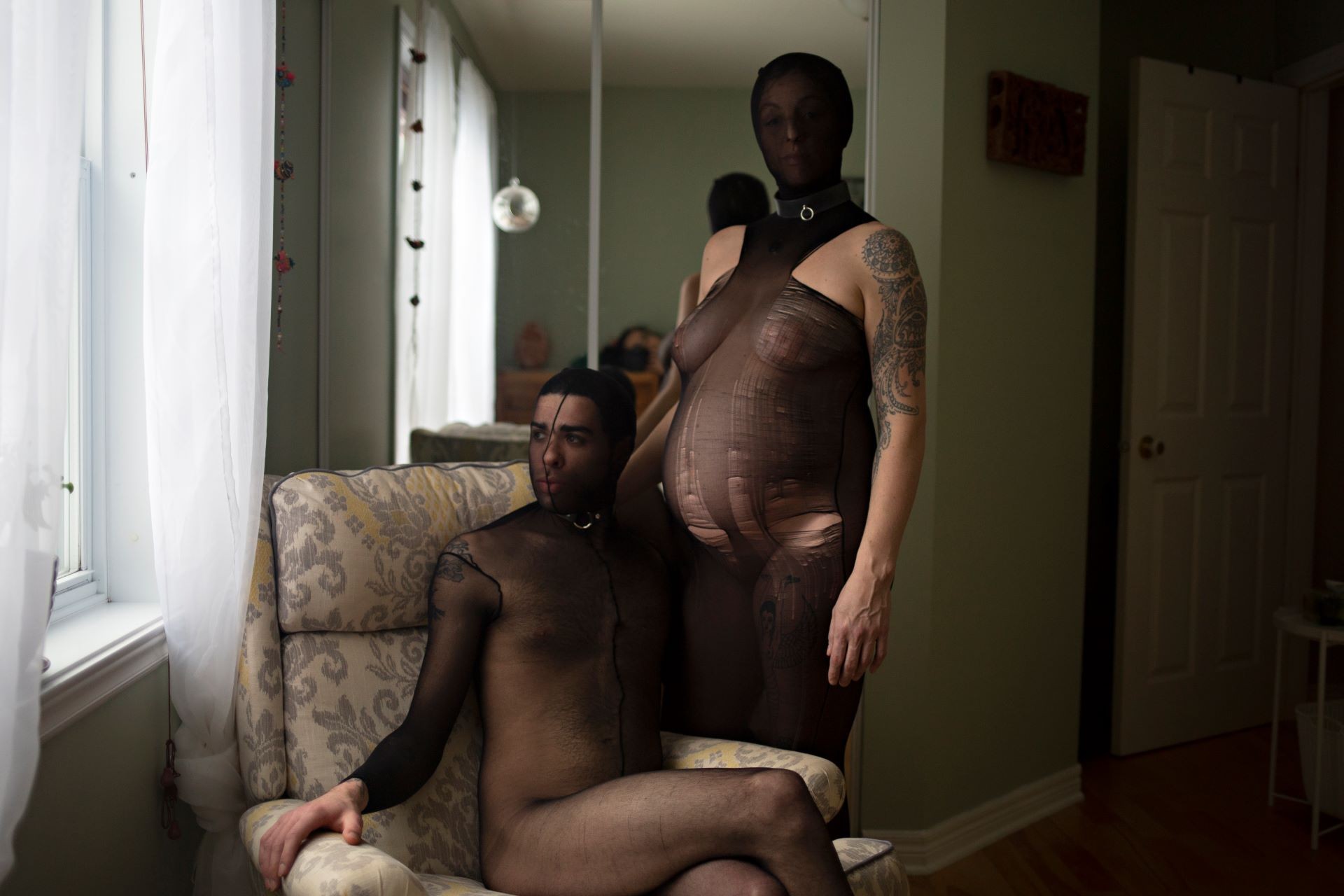 A queer couple posing in their living room dressed in a sheer black bdsm outfit with their face covered, one is pregnant and standing next to a male presenting companion sitting in a armchair