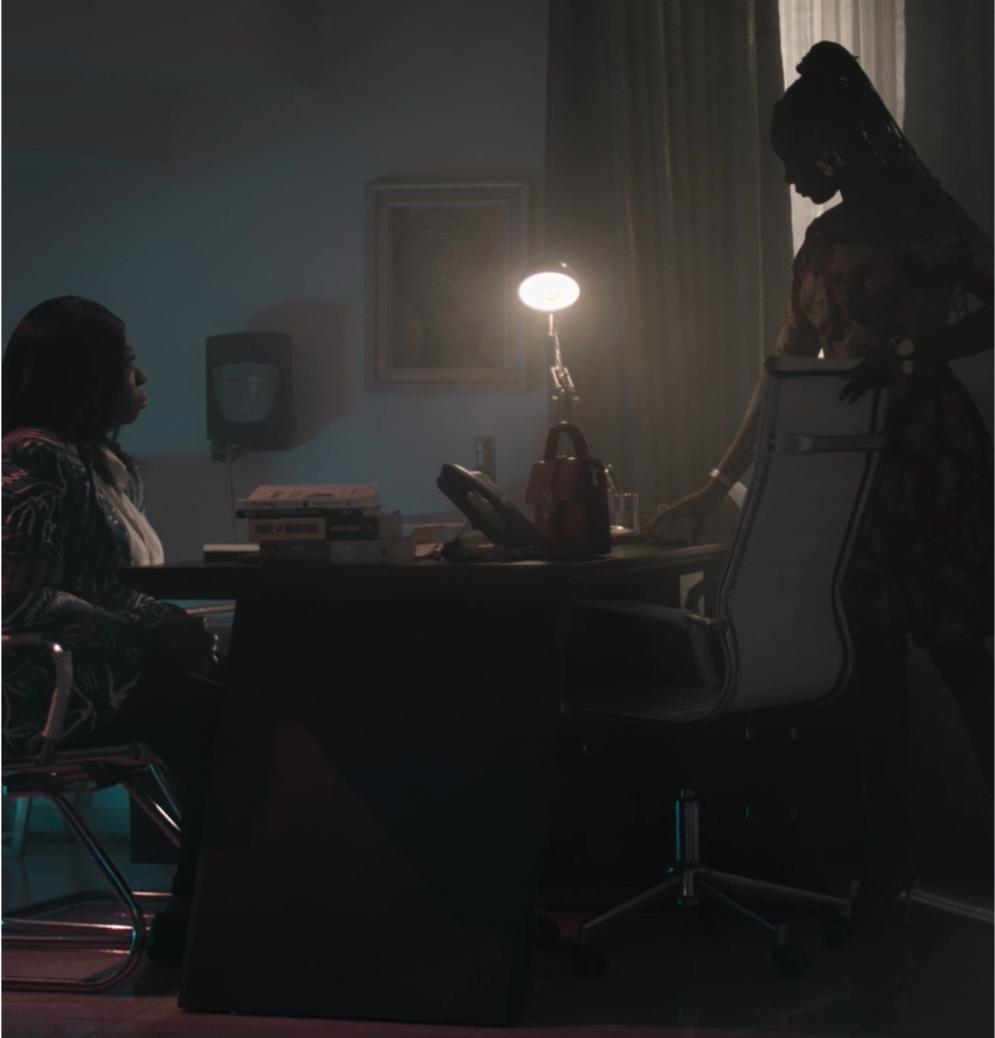 Two Black female bodied individuals are captured having a serious conversation in a dimly lit office. One of them is about 40 yrs old and sitting on a chair facing the younger protagonist who is standing on the other side of the desk, she looks like she is in her late teens. 