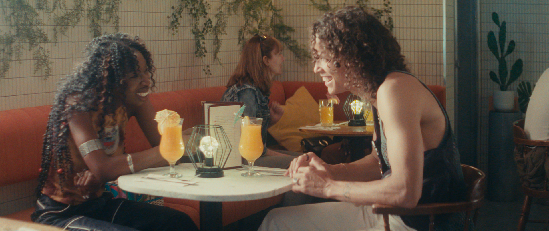 A Black woman is captured on a date with her estranged mixed-raced male identifying ex. They are conversing over two glasses of orange juice at a brunch restaurant.