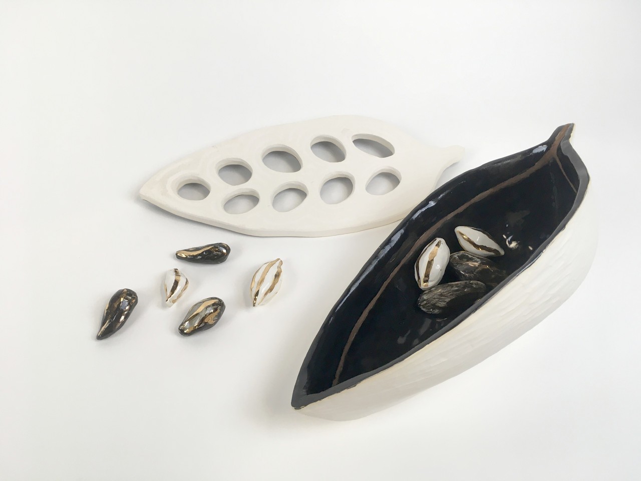 Picture of the full artwork. The documentation shows a small grouping of  small groupings of black and white glazed earthenware with gold luster displayed outside and inside the cacao bean. We can see the interior of the bean which is black with gold luster streaks and its white matte exterior. Along side the ceramic vessel there is a white matte ceramic leaf shaped piece with holes echoing the shape of the beans. 