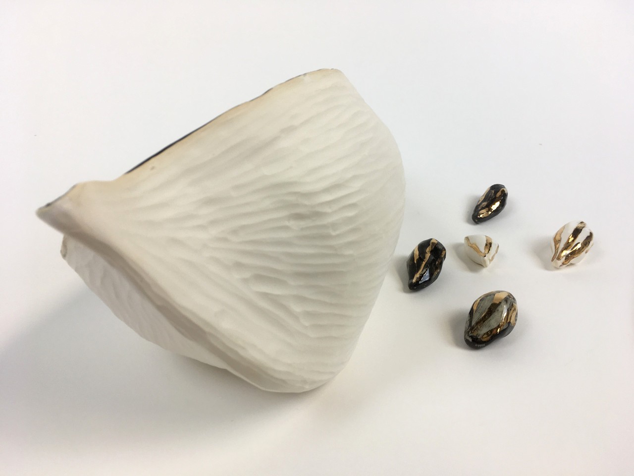 Detailed photograph of the work Seeds of Hope featuring a small grouping of black and white glazed earthenware with gold luster cacao seeds along side a rendering of the cacao bean. Only the back of the bean is captured, it is a textured white and matte earthenware. 
