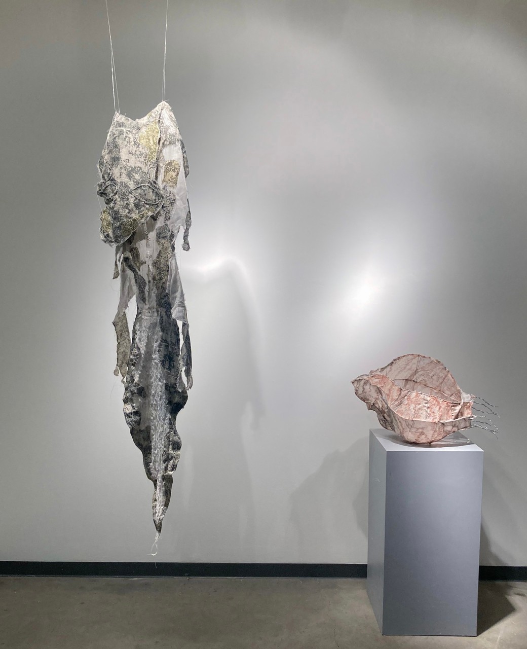 Installation view of the piece this was home which includes a peached colored rendering of a mollusk shell made out of metal wire and cheesecloth, as well as a green textile suit imitating shed lizard skin.    