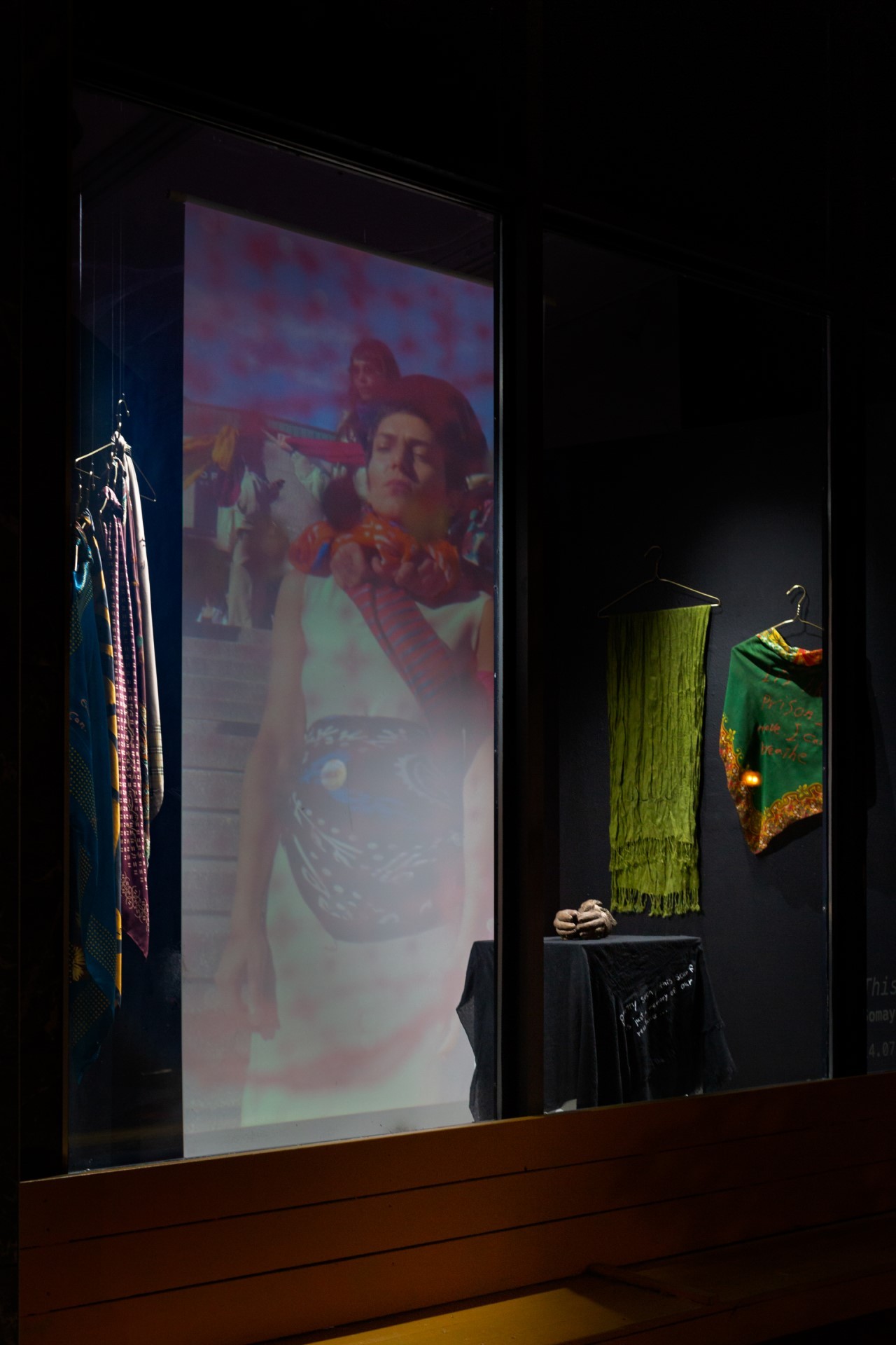 Documentation shot, prior installation of the work at La Central Power's front window. It showcases scarves hanging on the wall framing a projection of the artist performing and bronze casted hands displayed on a plinth. The hands are bound with a scarf. 