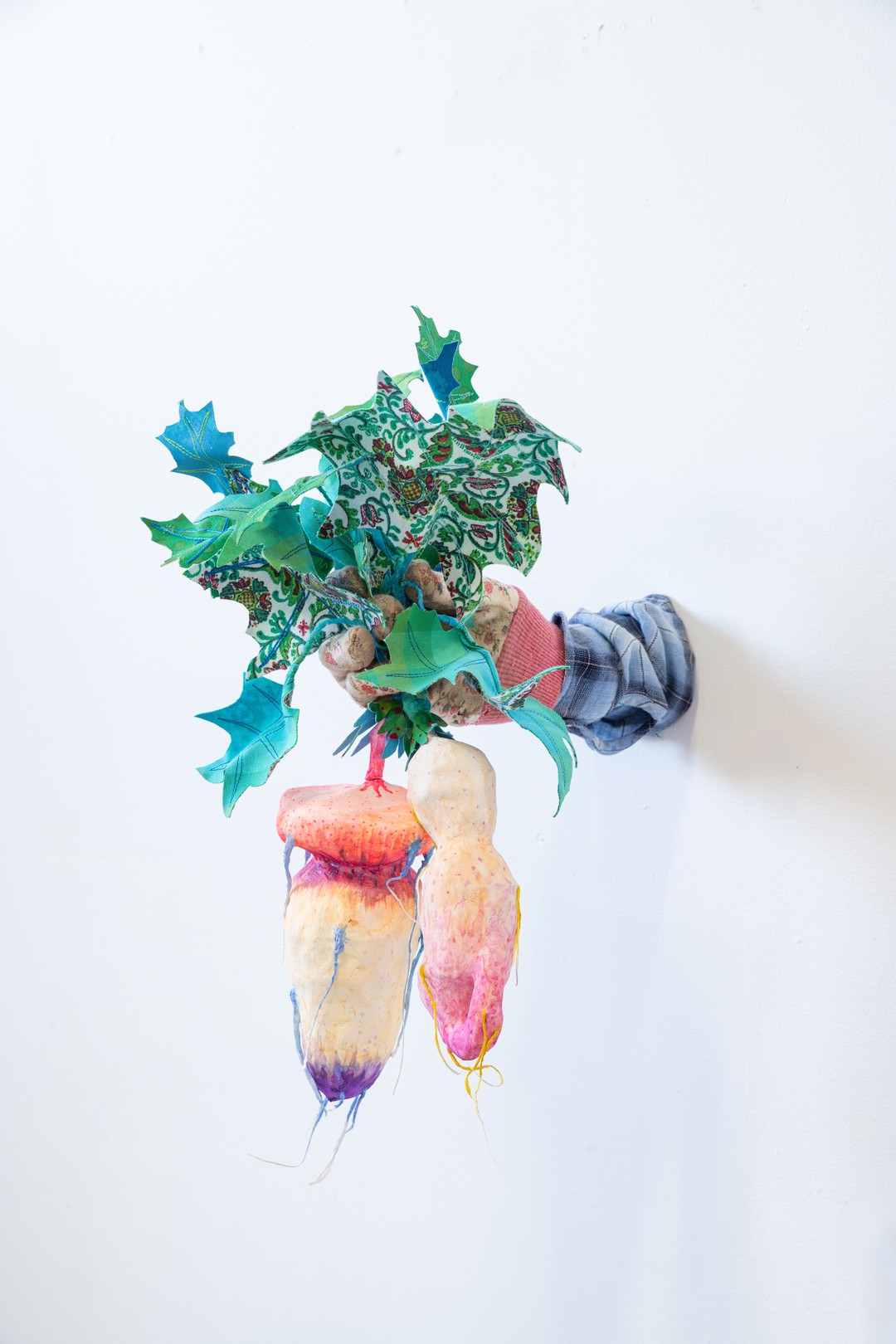 Sculpture in paper and textile depicting a hand holding a plant with green leaves and two white and flesh toned bulbs with phallic shapes.
