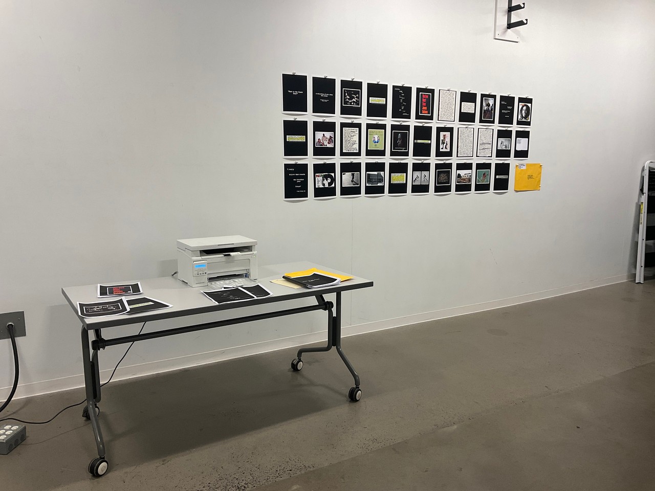 Installation shot showcasing printed archival materials mounted as grid on a white wall. A table alongside the grid has a printer and scattered archival materials.