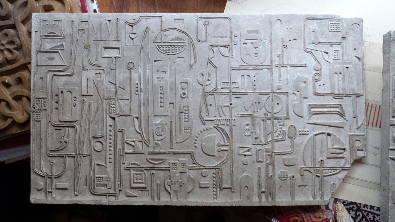 A photograph showing primarily a rectangular slab of concrete that has many geometric engravings. Visible at the edges of the photograph are several other materials, patterns, and papers from a studio archive.
