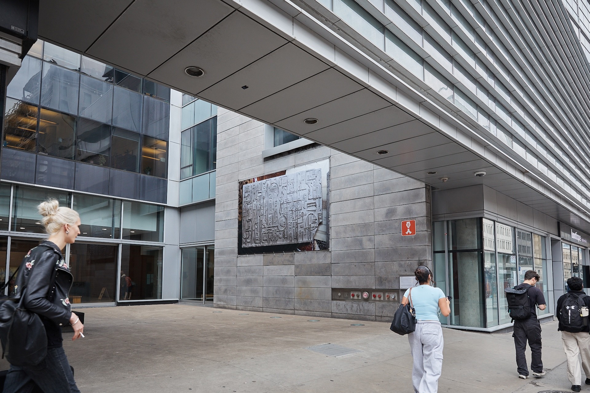 A large printed banner of a photograph is visible on the exterior of a cement and glass building. The image is taken from the street sidewalk. People are walking by from the left of the frame to the right. The wall that it is on contains a grid of horizontal rectangles. The printed photograph is of a model of a concrete relief. 