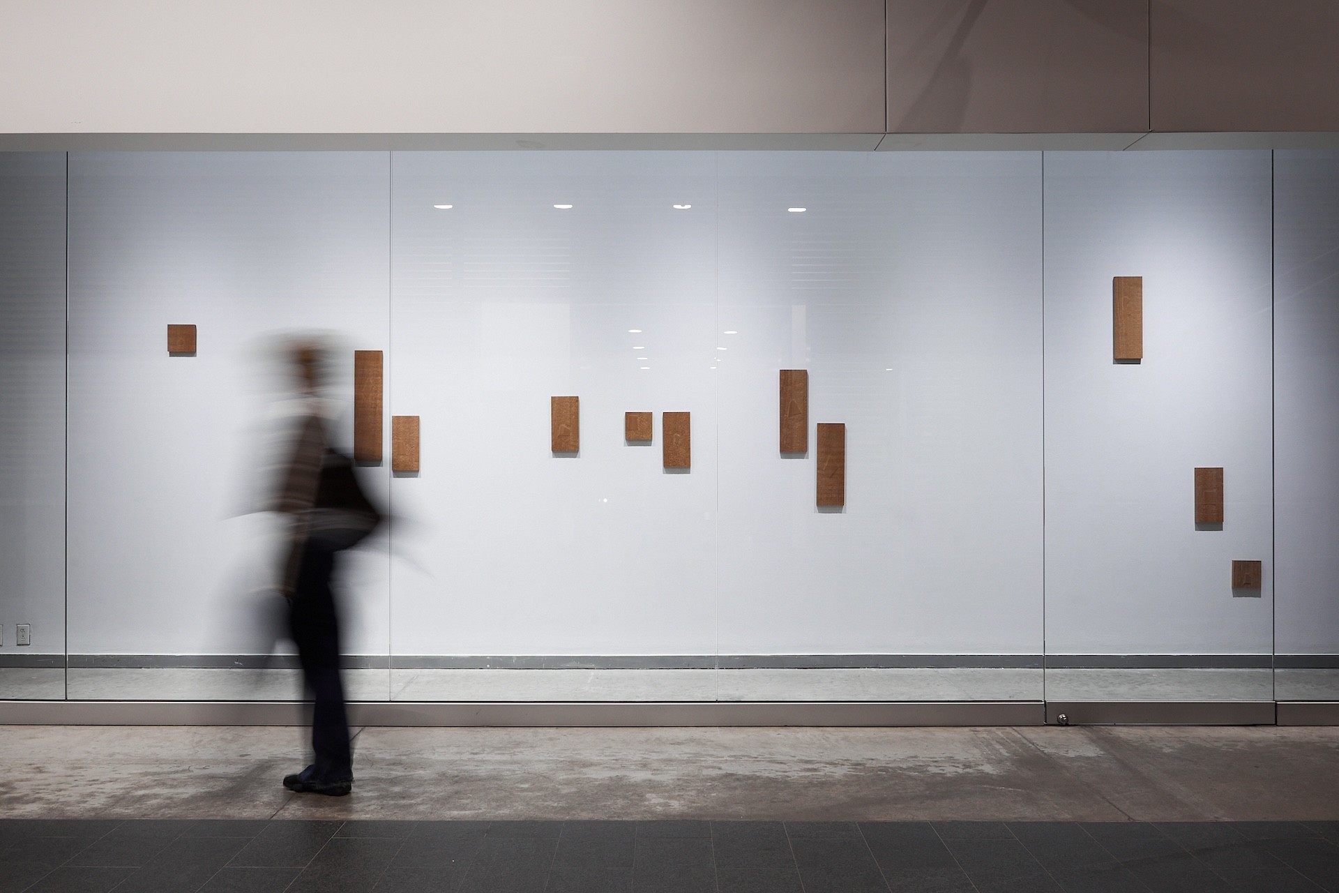 View of artworks installed along a vitrine. Behind the glass, a white wall is punctured by an array of wooden rectangular sculptures. To the left is a blur of someone walking by the vitrines 