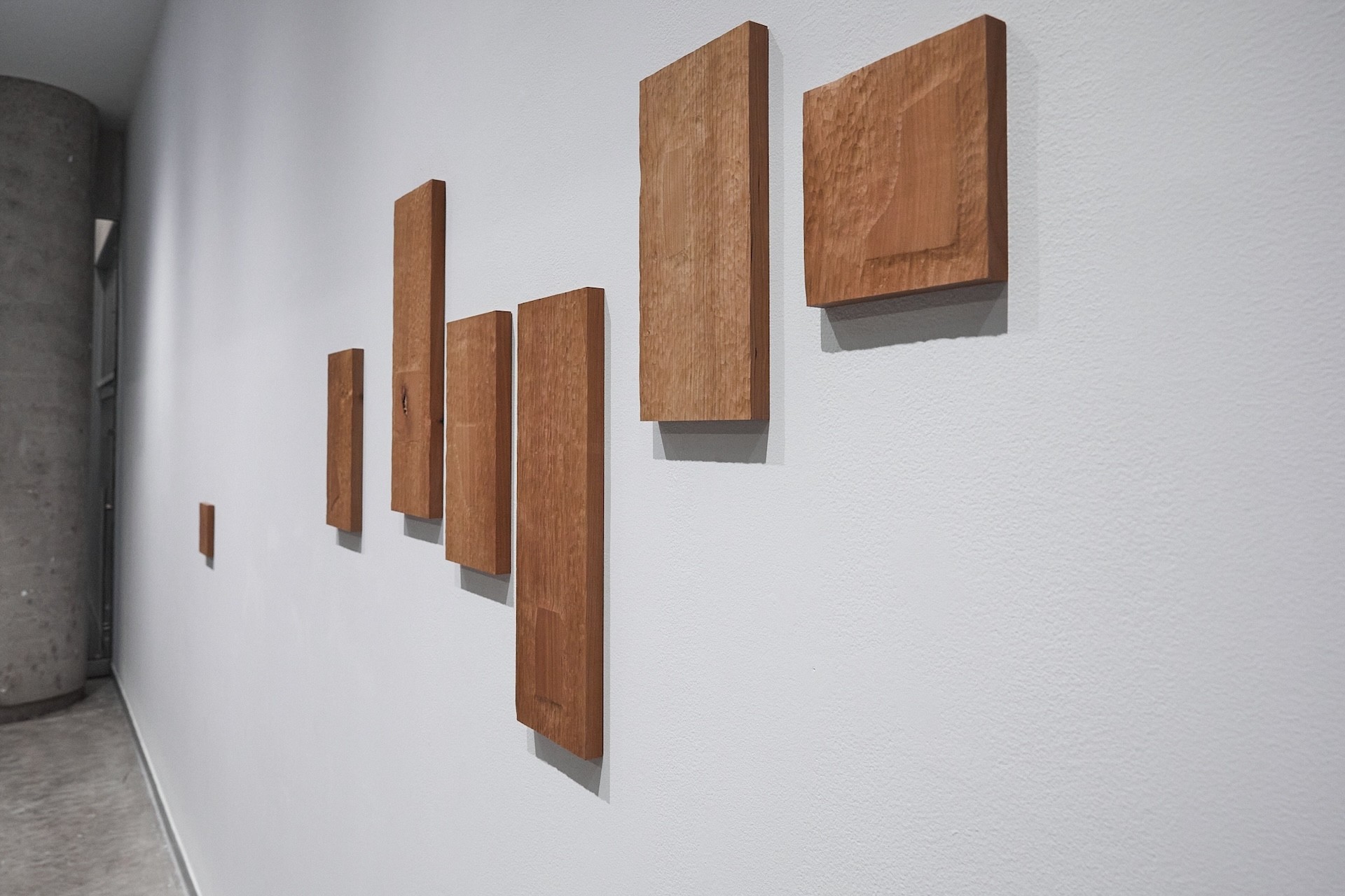Installation view of a white wall with several wooden sculptures installed. The pieces of wood are rectangular and show the texture made from repeated small gestures of carving. Each object has a shape that has the original flat wood finish, which shows the shape of a fallen mosaic tile. The vitrine is interrupted by a cement column.