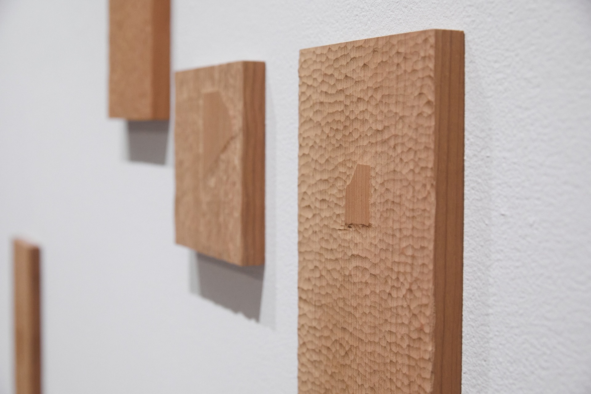 a close up view of a white wall with three wooden sculptures installed. The pieces of wood are rectangular and show the texture made from repeated small gestures of carving. Each object has a shape that has the original flat wood finish, which shows the shape of a fallen mosaic tile.