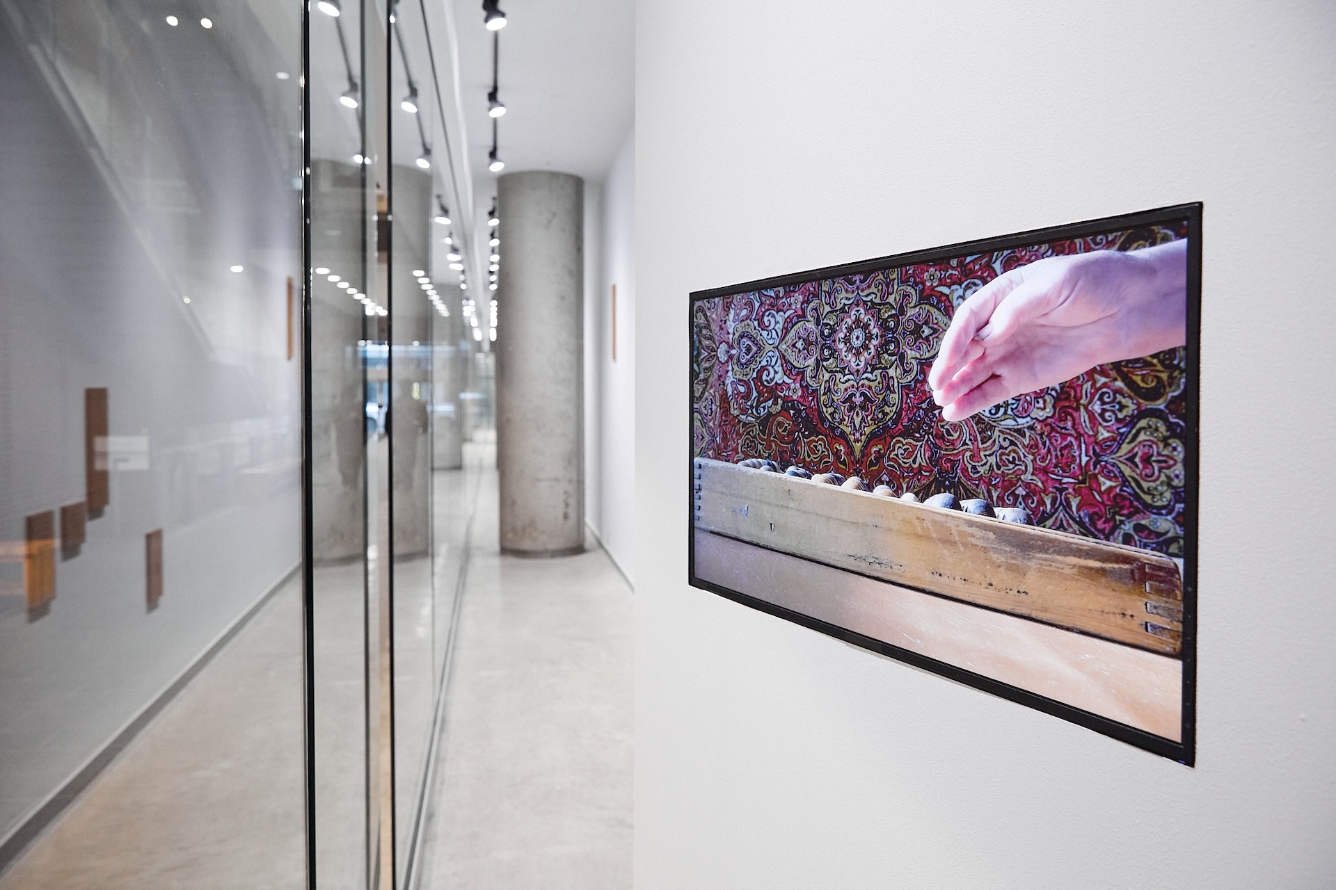 Close up view of the video installed in a corridor vitrine, on a white wall, angled differently than the vitrine. The video is frozen on a hand lifting towards or away from an abacus. The rich patterned textile that is the background of the video stands out amidst the whites and grays of the corridor and vitrines.