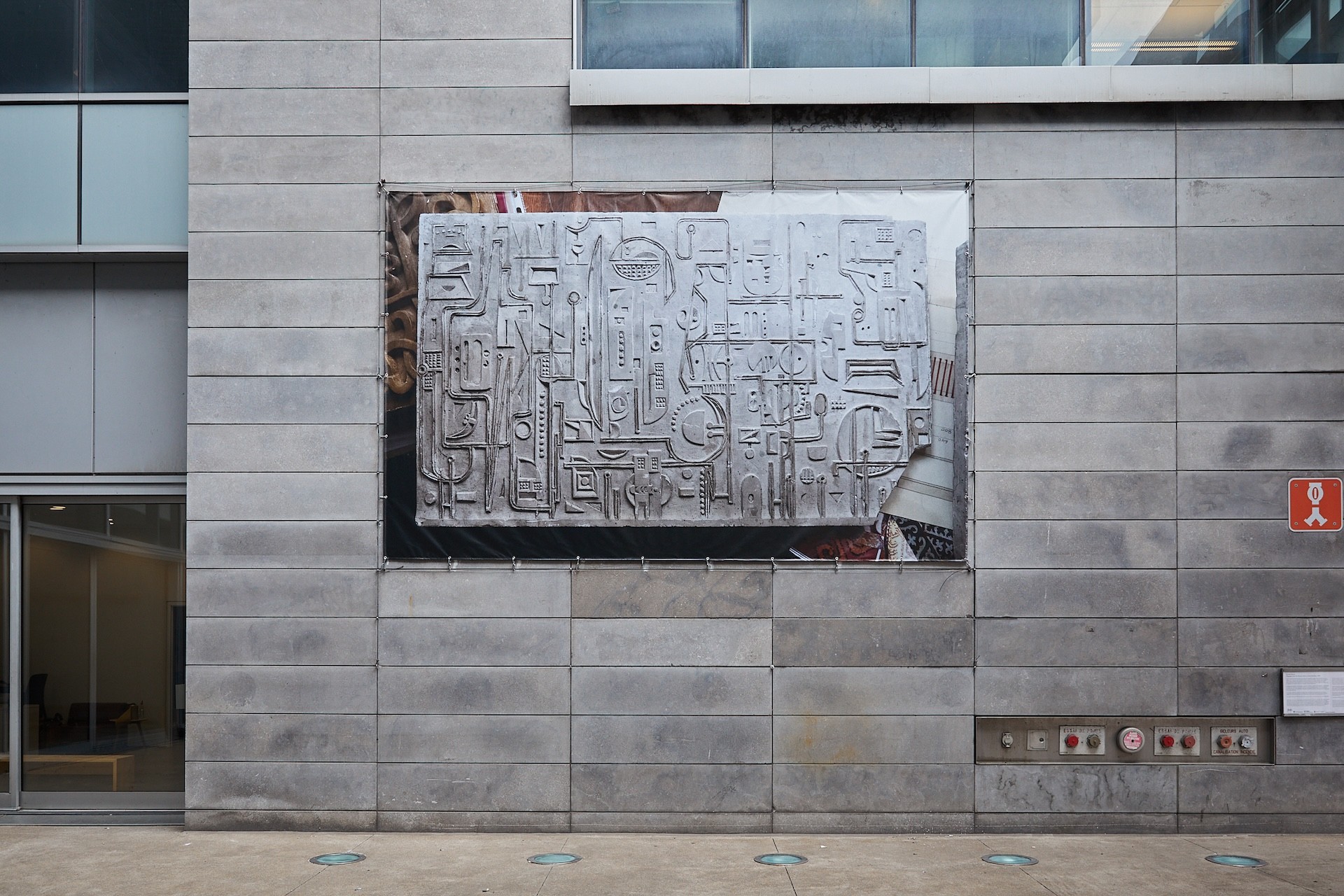 A large printed banner of a photograph is visible on the exterior of a cement and glass building. The wall that it is on contains a grid of horizontal rectangles. The printed photograph is of a model of a concrete relief, with other sculptural and paper items visible around the border of the object.