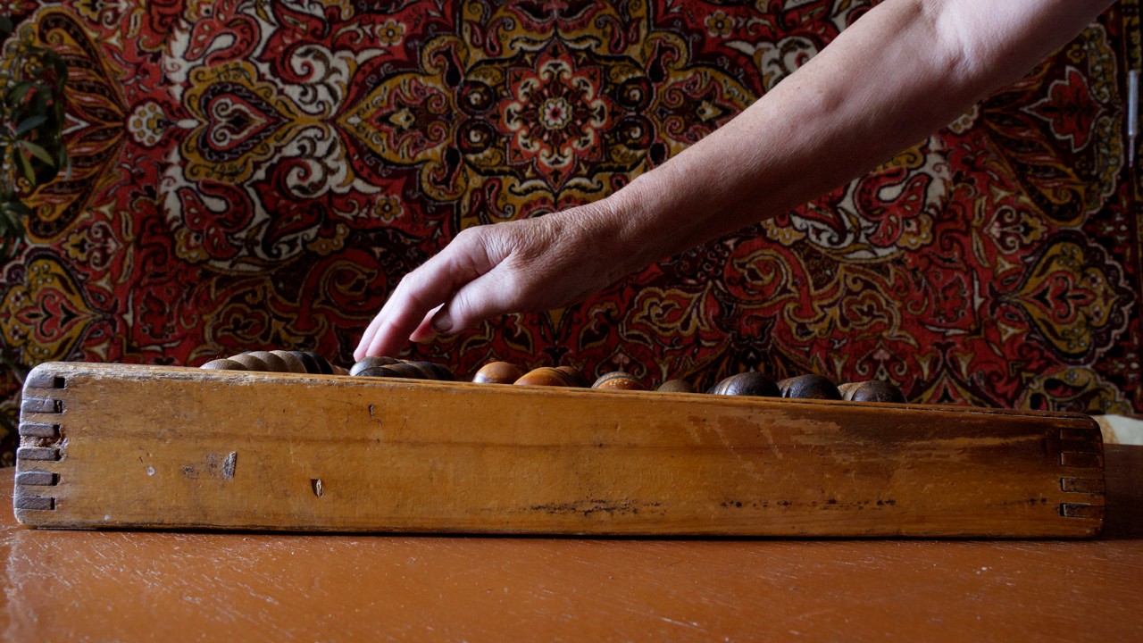 The video still shows, from the side, an abacus made of wood on a wooden table. A hand is reaching into the frame and touching the abacus beads. Behind the table and hand is a richly patterned textile serving as a backdrop to the image. The fabric is covered in patterns reminiscent of fillagree, in black, cream, red, and yellow colours.