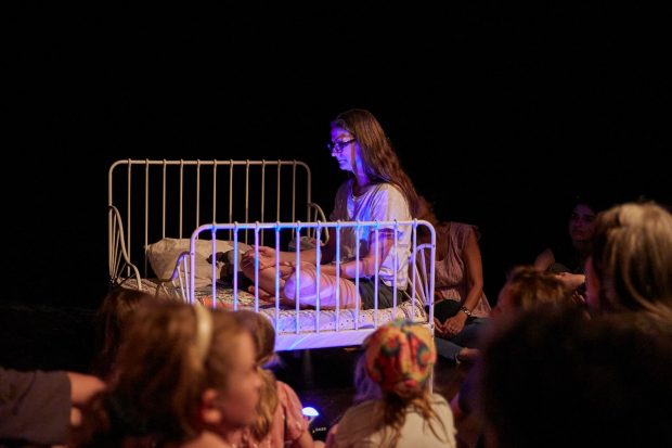 View of a white-walls gallery installation where man and a toddler are sitting in front of each other in front of a bed with two headsets to each side; a bunny lamp is next to the man