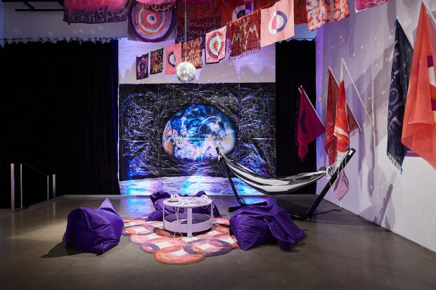 An art gallery installation of several flags hanging from ceiling supports with statements about mothernism. A tent is in the middle of the room, the ground is a purple carpet.  