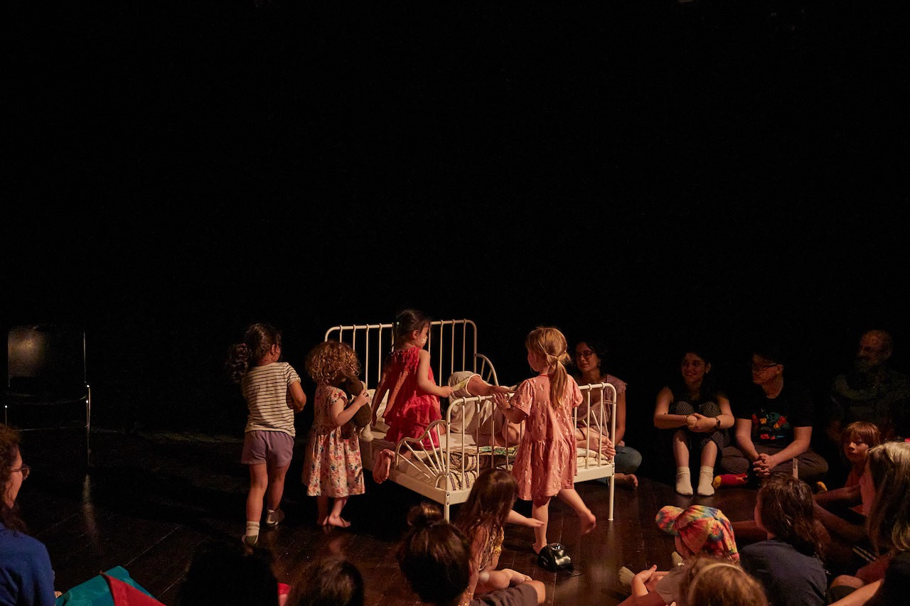 view of a black box gallery space with several young children sitting on or climbing into a small bed with metal head and base boards. The scene is illuminated in warm light in contrast to the darkness that the audience is in.