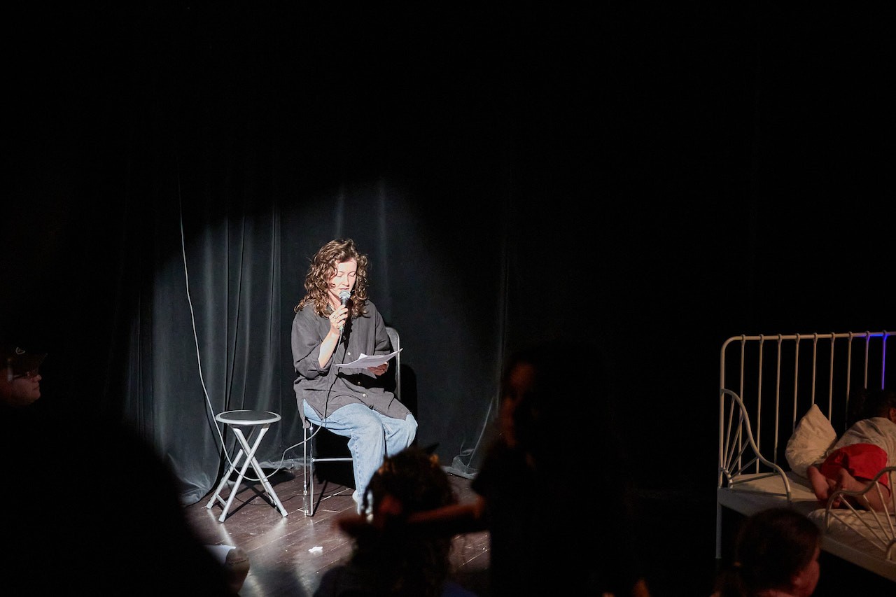 The performer Celeste Midori is reading from a printed paper, holding a mic and sitting on a chair. The room is dark, the curtain behind them is black, and a round spotlight highlights their performance.