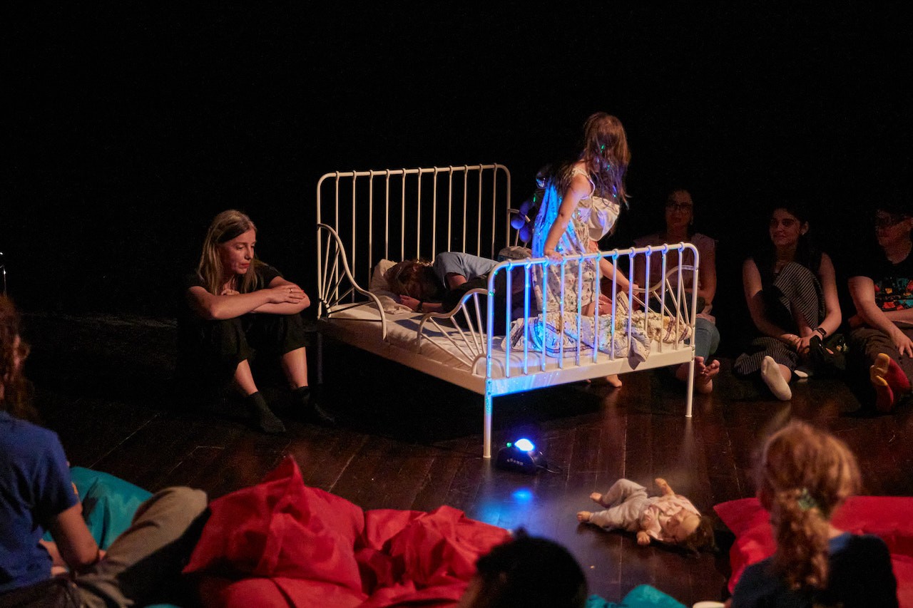 Performer Magdalena Olszanowski is sitting on the floor holding her knees bent to her chest. She looks pensive and quiet. To her left, children crawl, lie, and walk around a small bed. The scene is illuminated in the dark performance space.