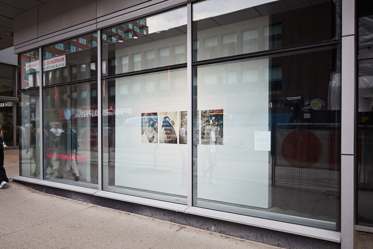 Exterior view of the FOFA Gallery vitrine from Ste Catherine street. The title of the exhibition "The Let Down Reflex" is written in pink on the bottom left section of the window. Some business signs are visible on the glass reflections. 