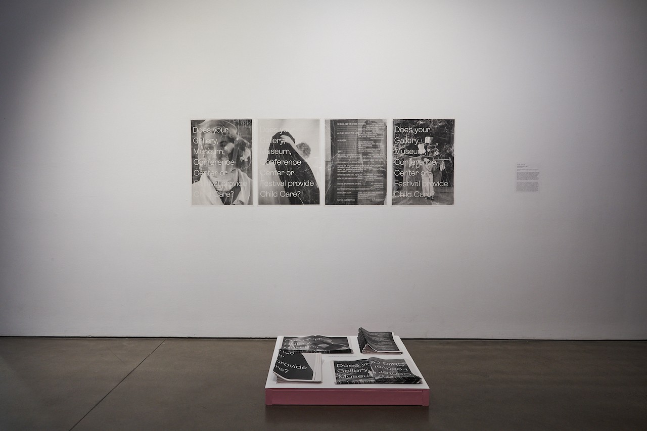A white gallery wall shows four black and white posters pasted in a row. On the floor in front of them is a low plinth with pink walls. On the plinth are piles of folded posters for attendees to take. 