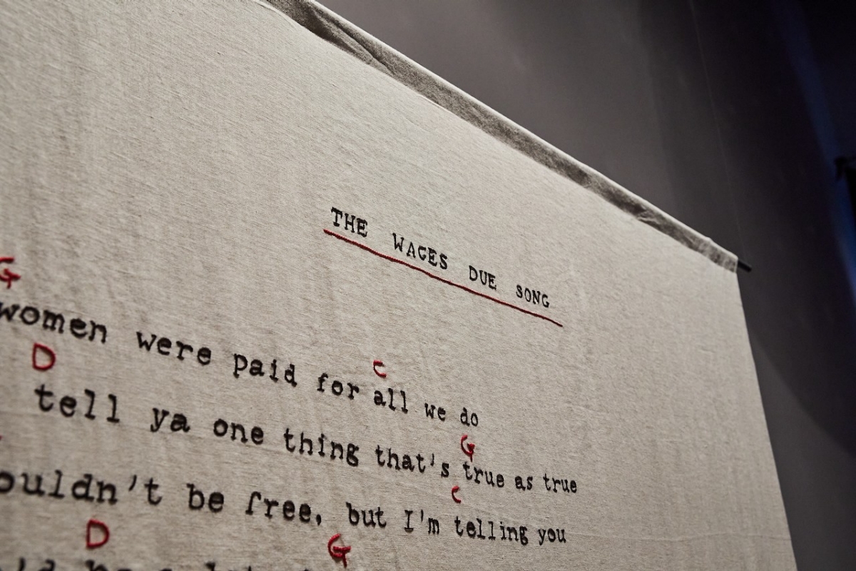 a photograph showing the details of a linen banner embroidered with song lyrics. The words are embroidered with black thread and the notes are embroidered between the lines in red. The linen is a creamy colour, similar to the aged archival paper it draws inspiration from. 