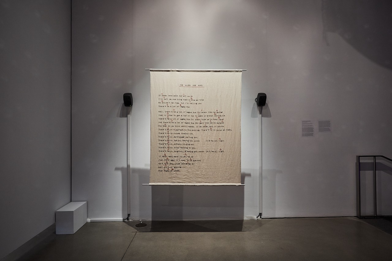 Installation photograph showing a large linen banner hanging from a rod about a foot away from a white wall. Song lyrics are embroidered in black with red notes indicated. Two speakers are visible on the wall to either side of the banner. 