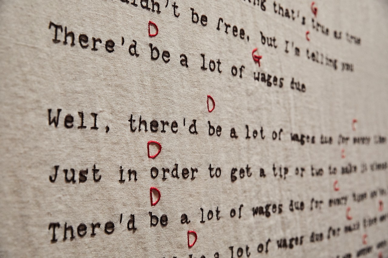 a photograph showing the details of a linen banner embroidered with song lyrics. The words are embroidered with black thread and the notes are embroidered between the lines in red. The linen is a creamy colour, similar to the aged archival paper it draws inspiration from. 