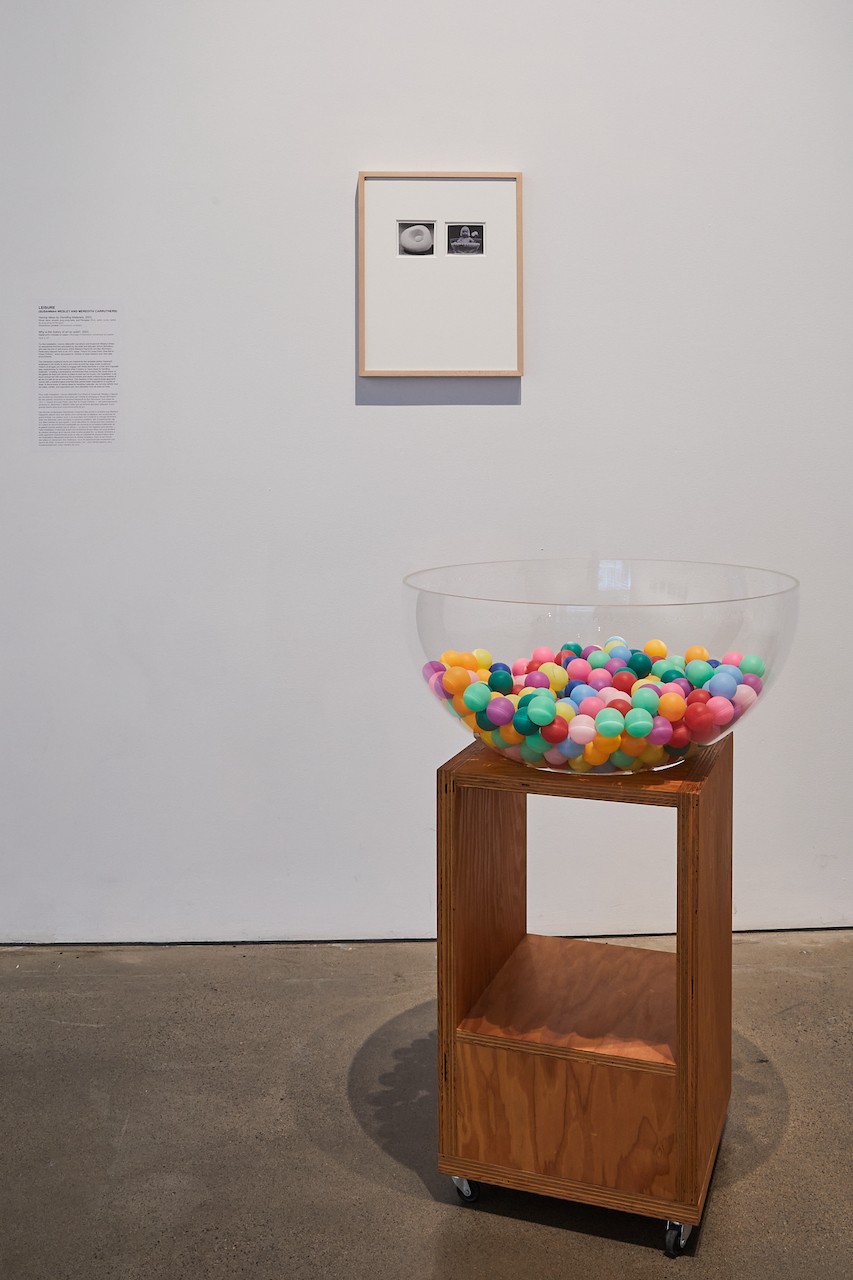 A gallery space with part of the Leisure installation visible. The white wall shows a framed piece with two images, one of a baby in a bowl and the other of a sculpture. There is a significant amount of white around the images, before the pale wooden frame. In front is one wooden sculpture, with a large transparent plexiglass bowl on top, and dozens of colourful pingpong balls inside. 