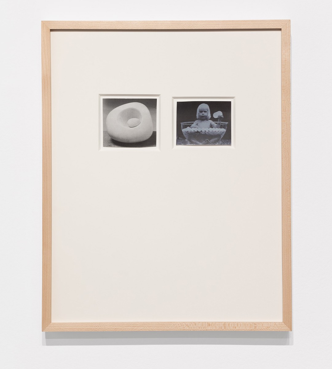 A white wall with an artwork hanging on it. The piece is framed in light wood and has two smaller images within it. One is a rounded sculptural pale shape the other is a baby sitting in a bowl of water with pingpong balls.
