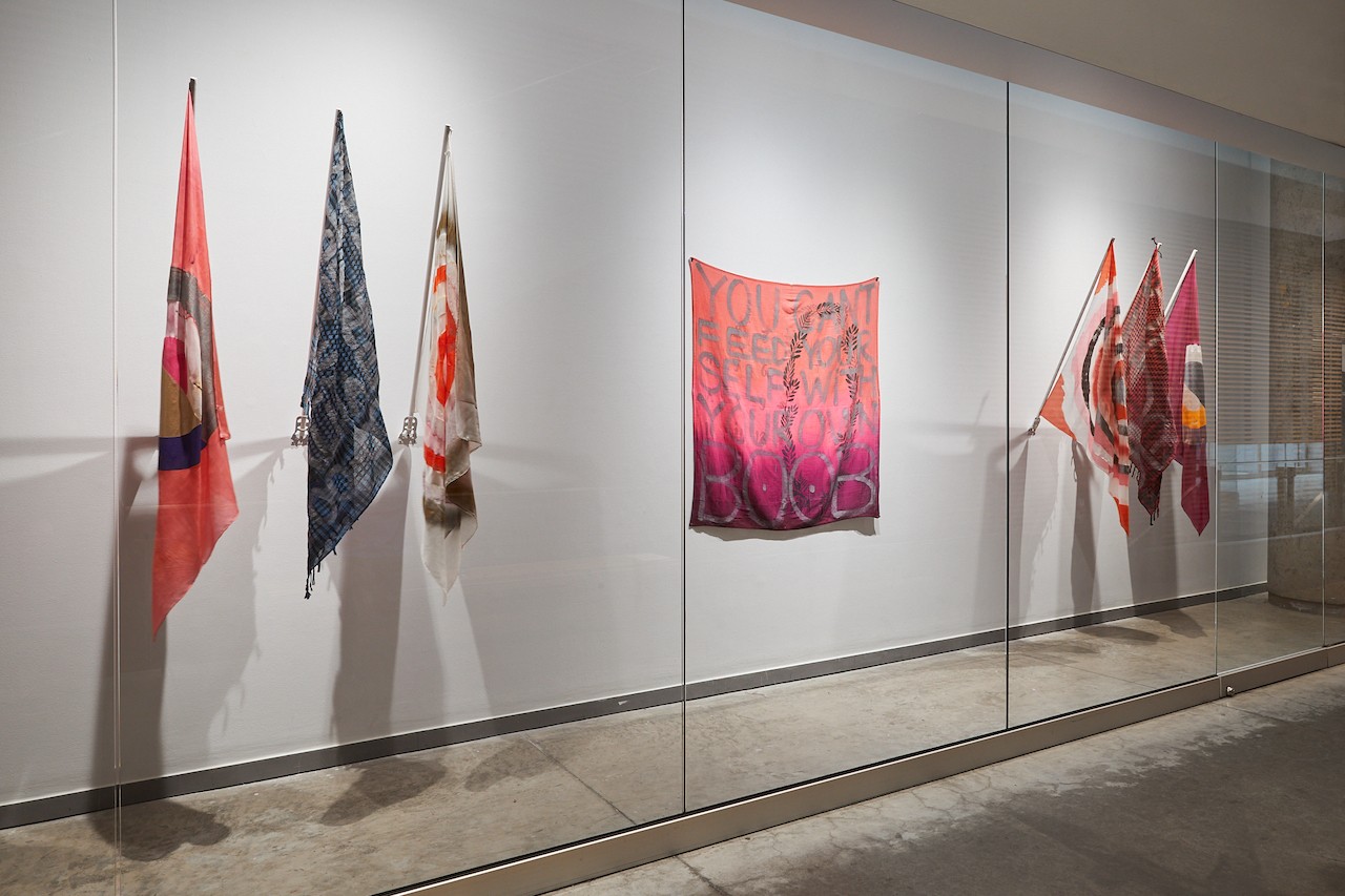 A corridor vitrine with two sets of three hanging flags. Two of them have grid patterns, and all are painting on warm colours, like orange, dark red, magenta, and brown. The details of each design is not visible since they are hanging down. Between the two sets of flags is a square textile visibly open on the wall which reads "you can't feed yourself with your own BOOB" 