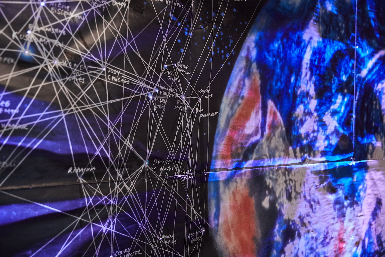 A close up of the large glowing blue planet earth print. The colour surrounding it, the black outerspace, is filled with names and lines that are drawn with silver ink. Glowing stars connect the various points.