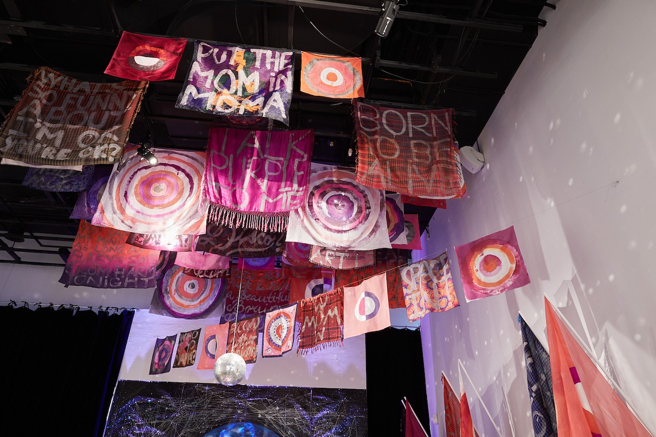 Looking up to the gallery's cieling, about a dozen flags with various designs and patterns hang from the ceiling. A disco ball hangs from the center and throws reflections upon the walls.