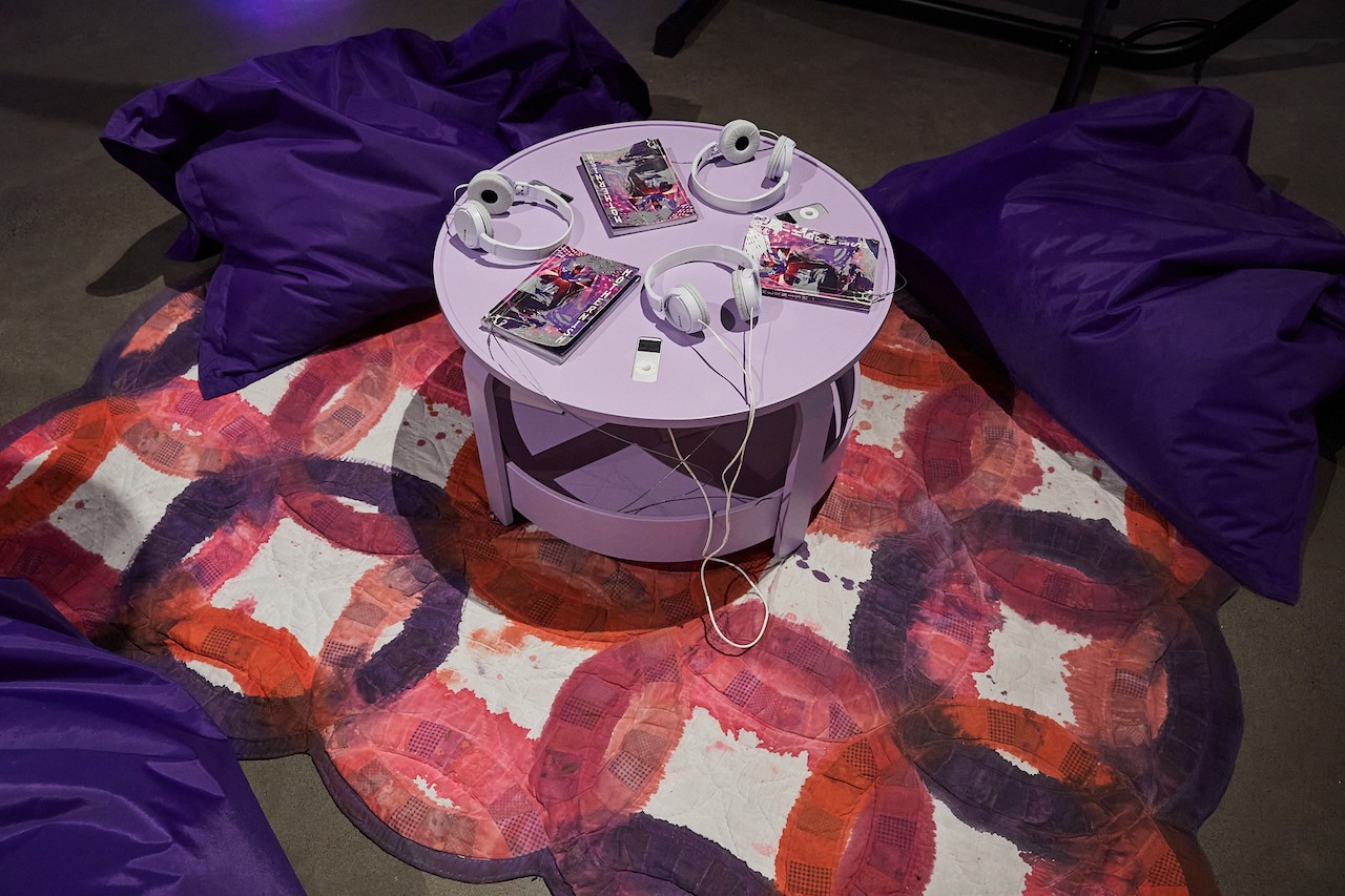 This image looks down at the gallery isntallation consisting of a fabric on the floor, dyed with rings of purple, red, and orange, a lilac-coloured round table, and three purple sandbags for sitting. Atop the table are three "mothernism" books, and three lilac-coloured ipods embedded in the table, each with their own headsets. The space looks comfortable and inviting.