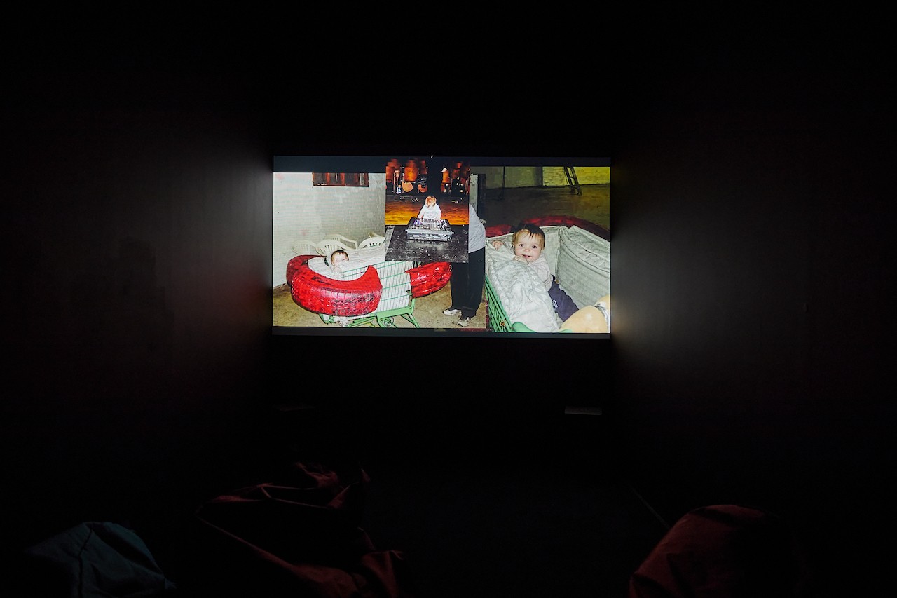A projection glowing in a black box room that shows a still from LoVid's video. The still is a collage of three photographs of a baby in a noise show venue. The baby is in a shopping cart, behind musical equipment, and in a crib. The images seem dated in their quality and aer