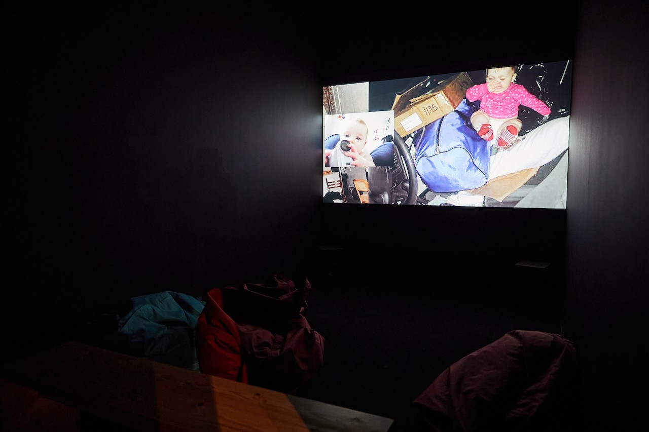A black box room is illuminated by a bright projected video. Some bean bag seating is dimly visible, occupying the ground of the room. The video still is primarily a baby surrounded by bags in the front seat of a car. Overlayed on this is a closer portrait of what seems to be the same baby. 