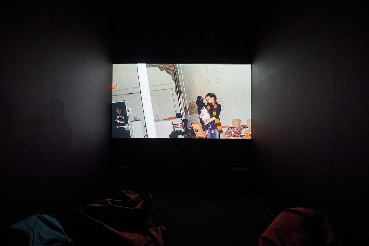 A black box room is illuminated by a bright projected video. The folds of bean bags in front of the projection reflect some of the video's light. The video shows a few people standing in a venue, one of which is smiling, has a long striped scarf, and is holding a baby that has hearing protection on.