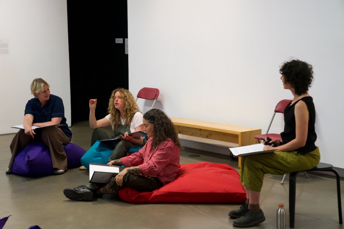 View of three pregnant performers sitting on large beanbags in a gallery space. They each have the script open in front of them. Rachel is sitting on a stool looking over at them. One performer is frozen in speech with her hand raised for emphasis.