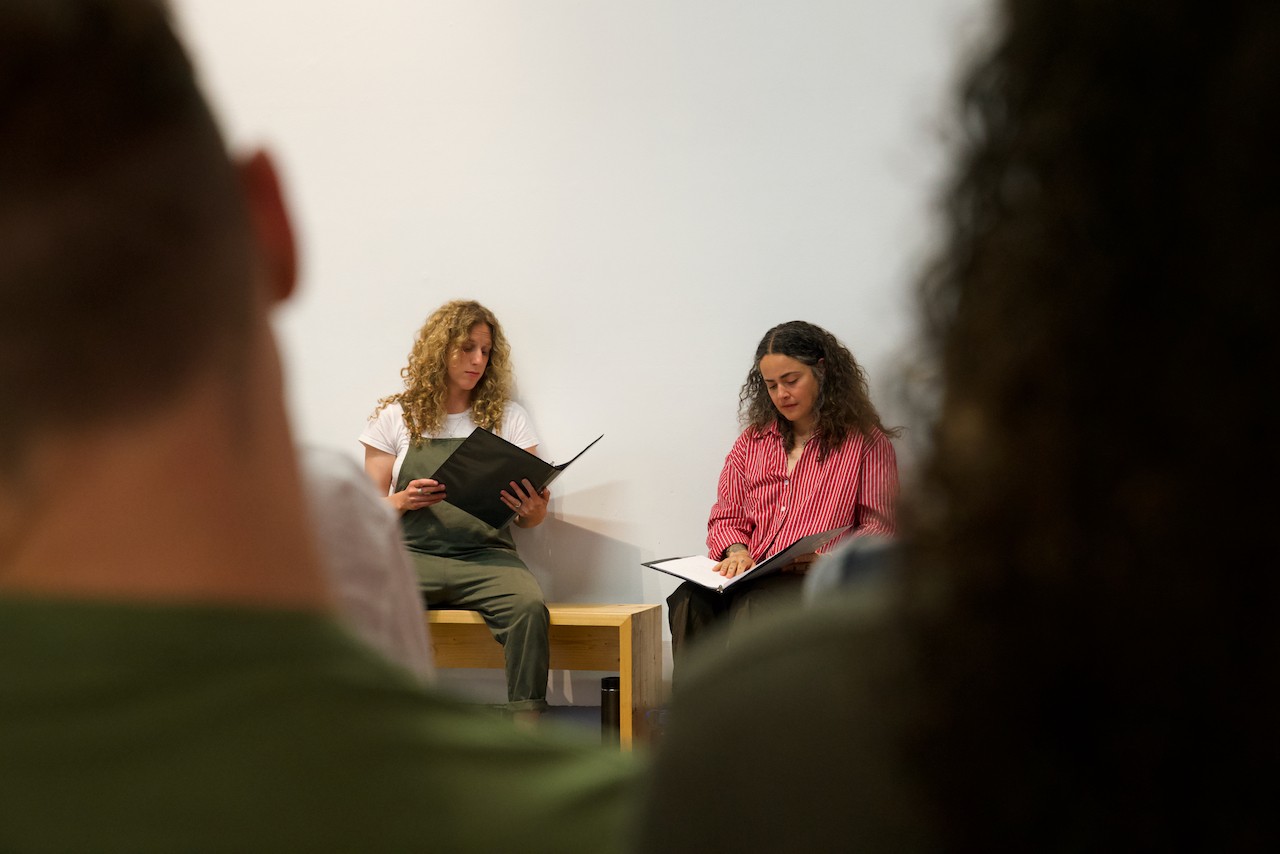 Two performers are visible sitting and looking down at their lines. The scene is visible between the outp-of-focus silhouettes of audience members in the foreground. Two heads flank either side of the composition. 