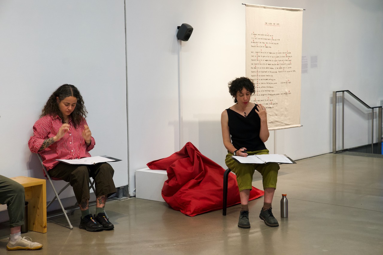 Two people are visible sitting in the gallery, reading their lines, speaking, and using their hands to emphasize. A large beige textile that is hanging some ways behind Rachel. It clearly has words embroidered on it, although they are not legible. 