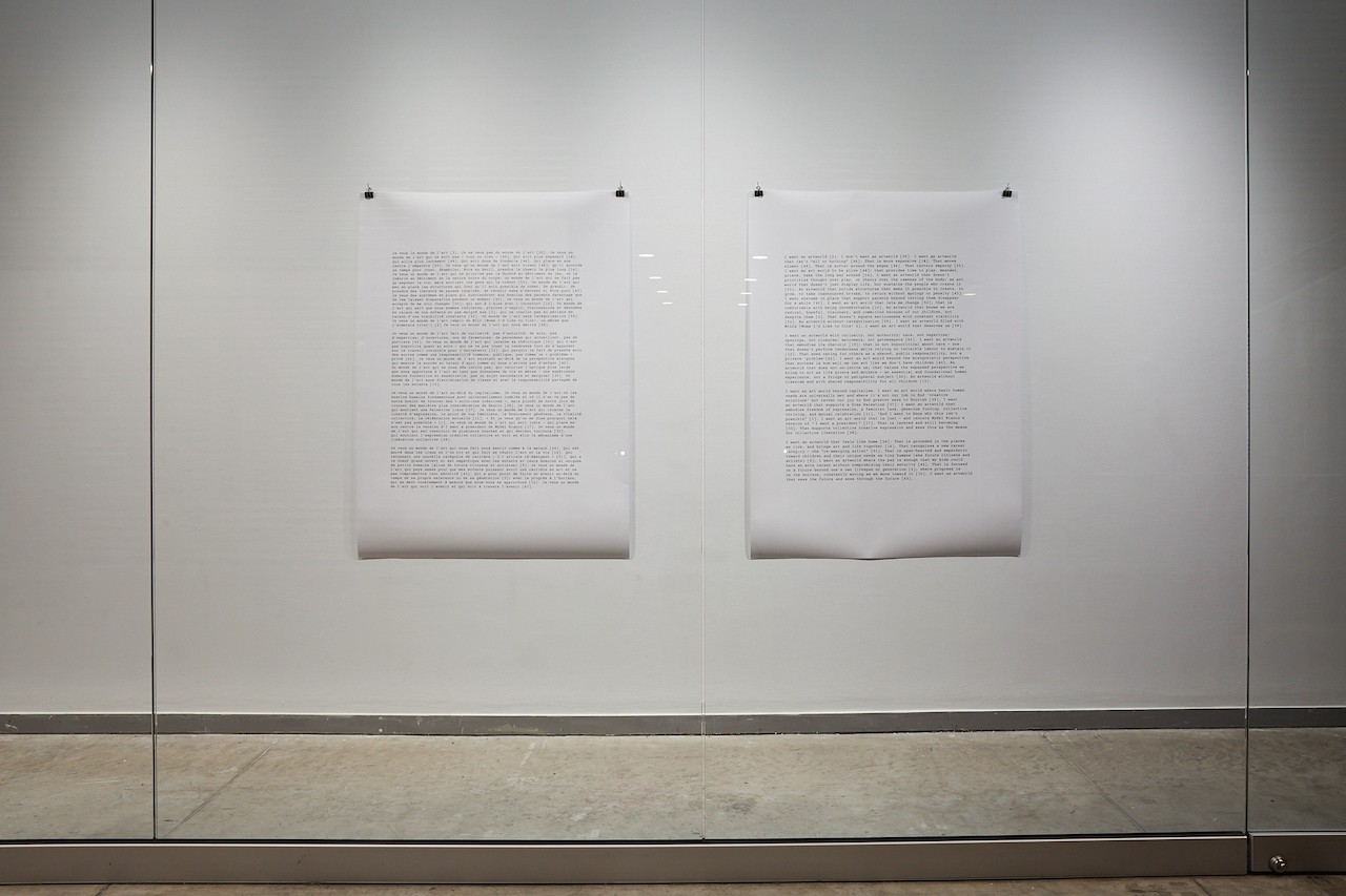 Two large prints are hanging in a corridor vitrine. Each print is of black text on white paper. The installation photo is not close enough for the writing to be legible.