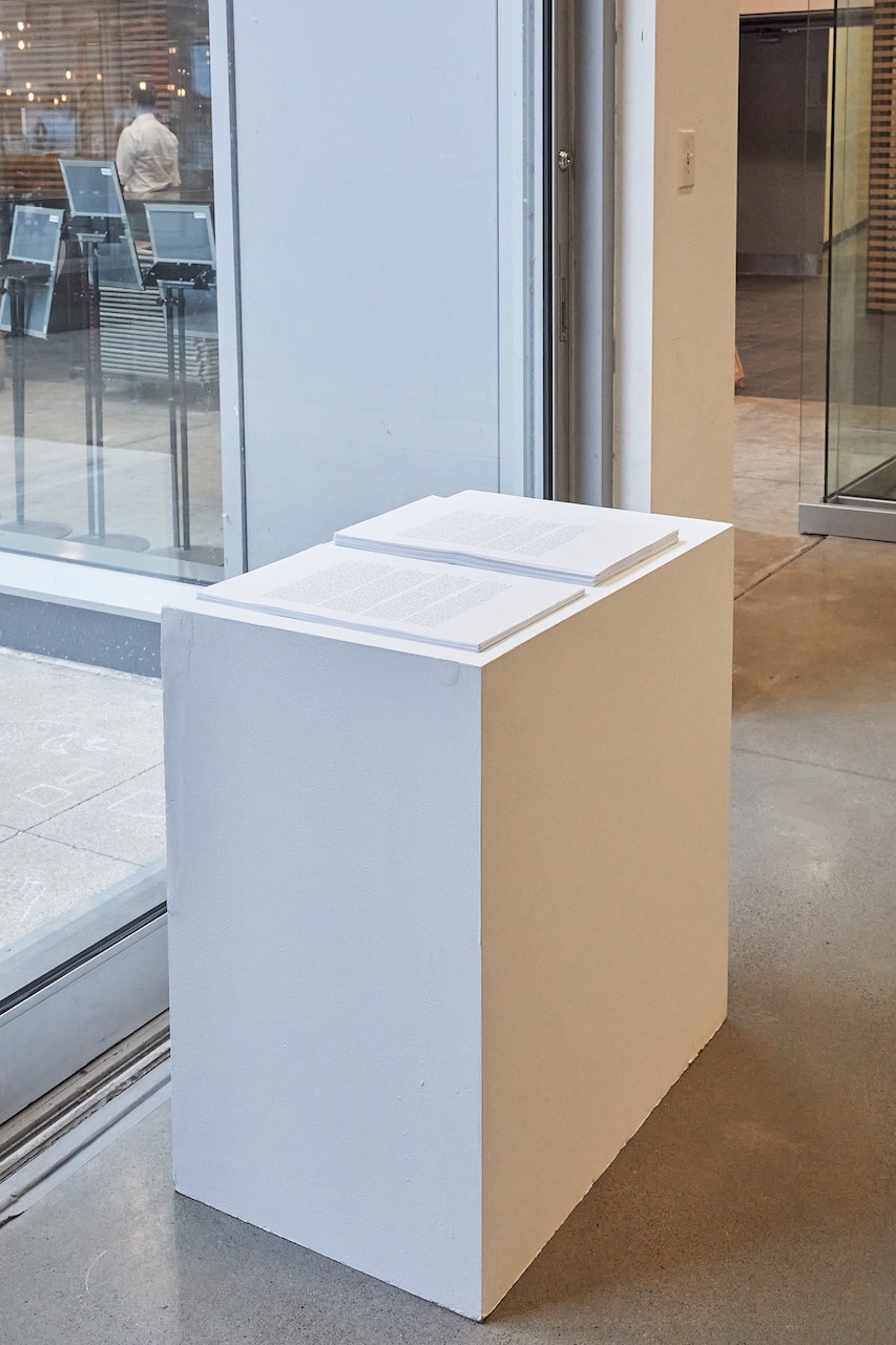 Inside a gallery space, a plinth is standing next to the entrance and in front of a window. On the white plinth are two stacks of papers, one that is the English text and one that is the French. They are clearly available to be taken.