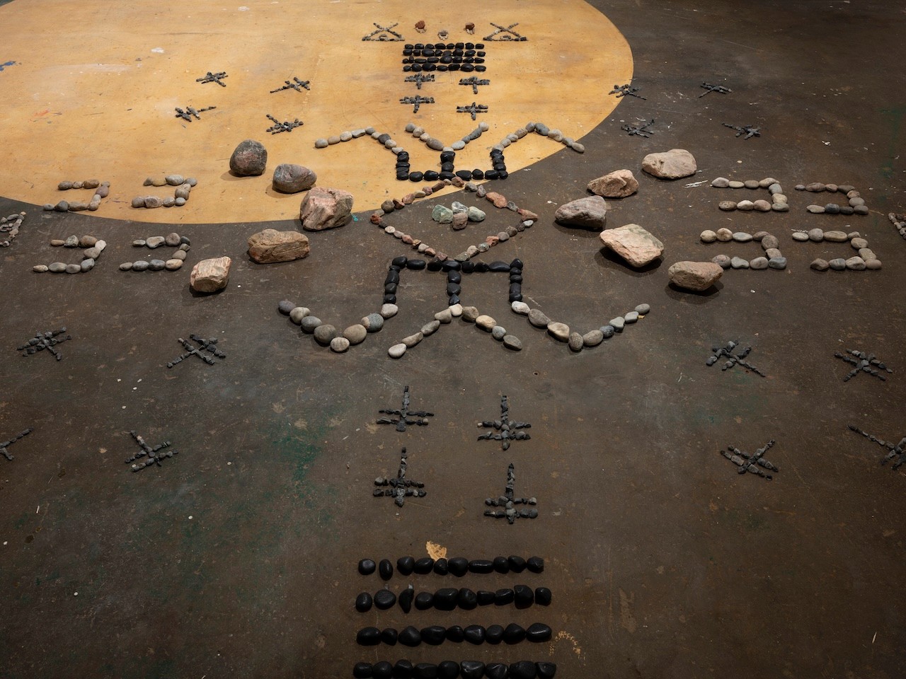 Photograph of a brown floor with a large yellow ochre circle. Radiating out from the center of the frame, rocks of different sizes create geometric shapes and patterns.
