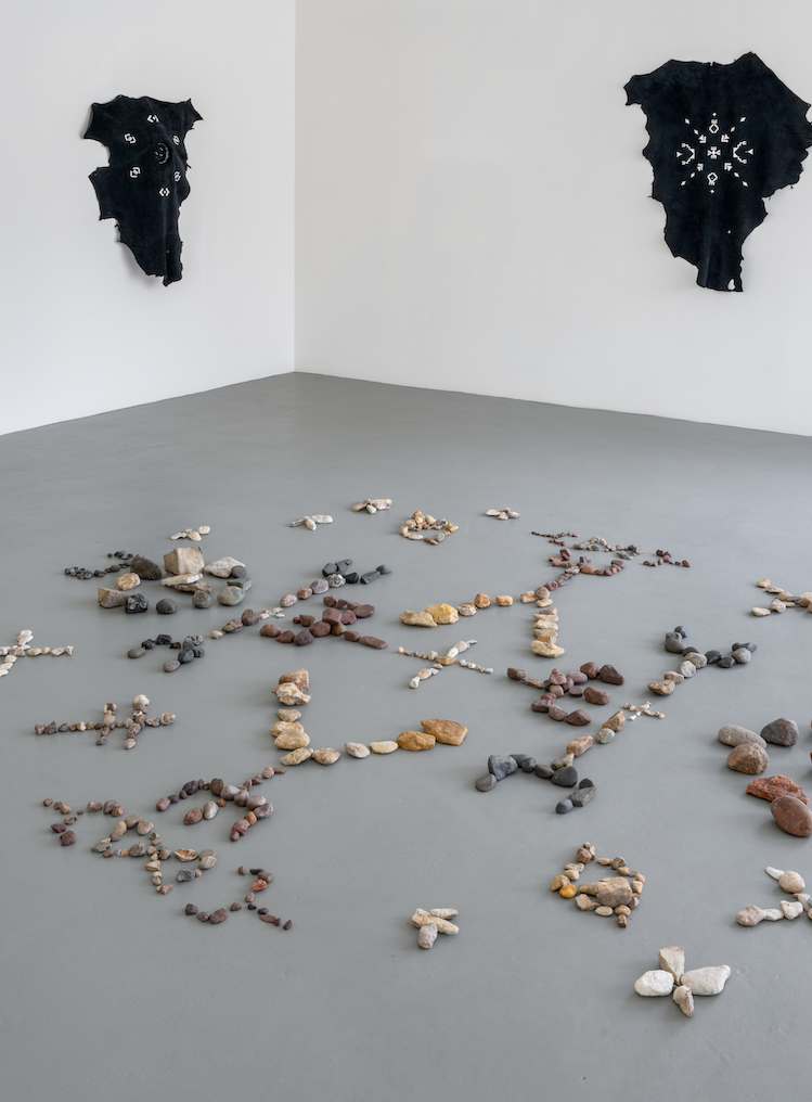 On the grey floor of a white cube gallery space is an installation of rocks forming geometric shapes. On the walls are two hanging embroidered hides.