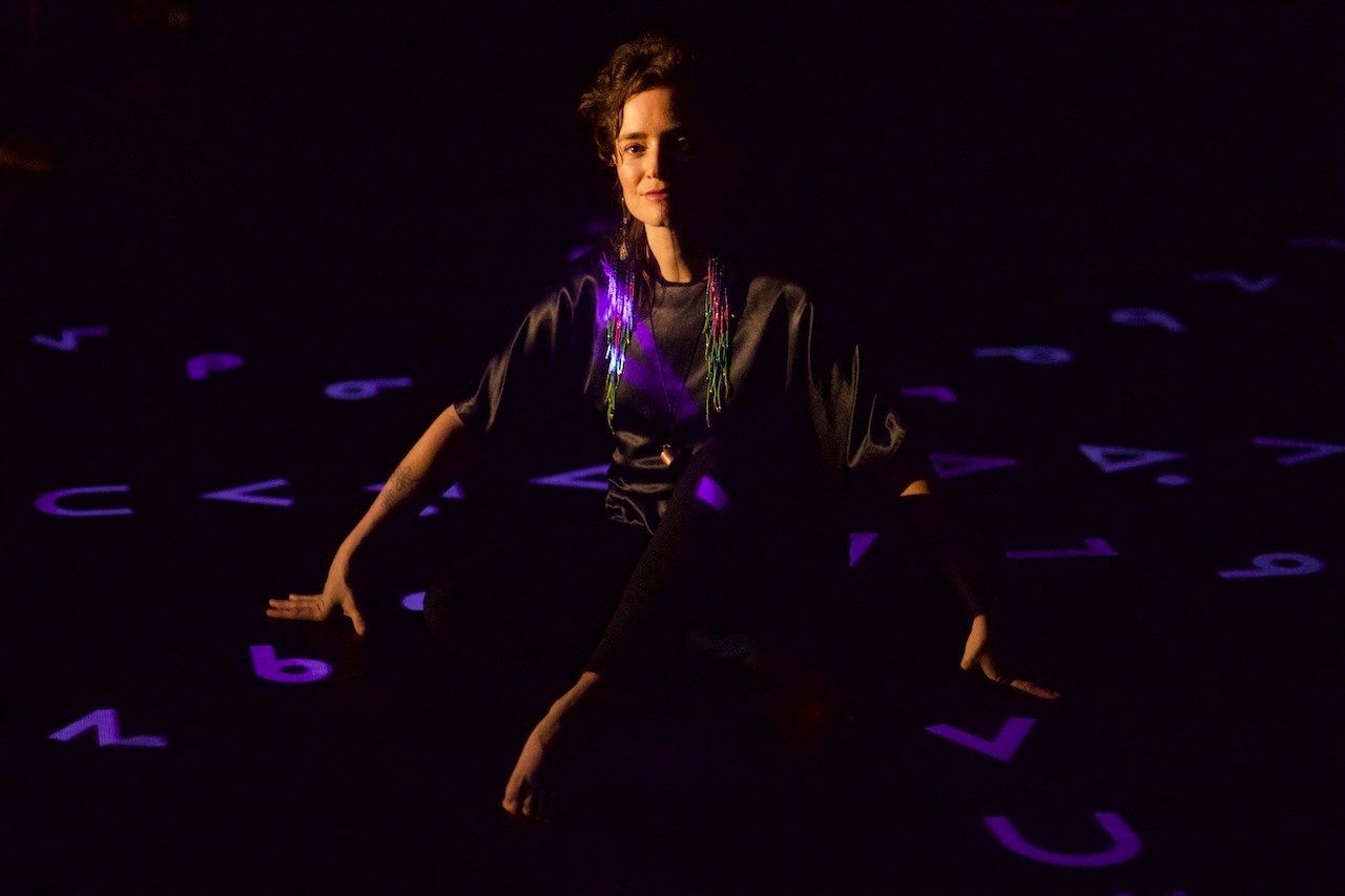 Moe clark sitting on the floor of a dark room with purple projections of cree lettering upon them and the floor around them. Their hands are perched on the ground to hold themselves upright