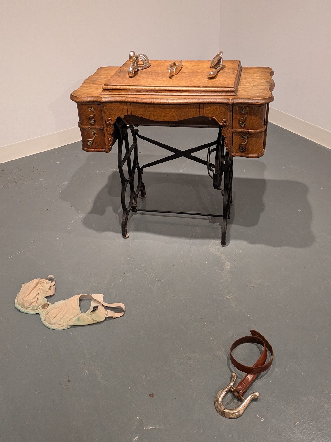 Ornate sewing table with three bronze objects laid on its surface and on the floor: a stained bra and a belt with an oversize buckle